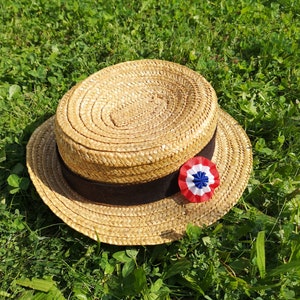 May include: A straw boater hat with a dark brown band and a tricolor rosette. The hat is made of woven straw and sits on a bed of green grass. The rosette features red, white, and blue colors.