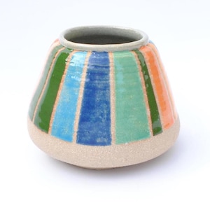Puede incluir: Un jarrón de cerámica con forma redondeada y boca ancha. Está decorado con rayas verticales en verde, azul, naranja y turquesa. La base es de color beige claro y el borde es gris. Un objeto decorativo.