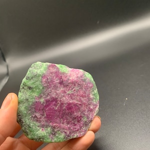 May include: A green and pink zoisite gemstone with a rough, natural texture. The stone is a light green color with pink and purple inclusions.