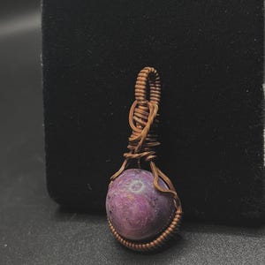 May include: A purple gemstone pendant wrapped in copper wire. The pendant is a round shape and has a wire loop at the top for hanging.