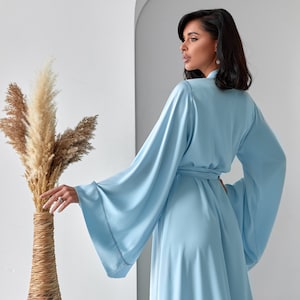 May include: A light blue robe with wide sleeves and a matching belt. The robe is made of a soft, flowing fabric. The model is wearing the robe. The background includes a tall vase with dried plants.
