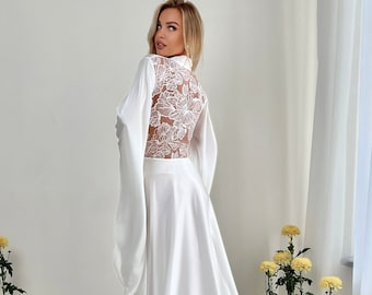 May include: A white satin wedding dress with long sleeves and a floral lace back detail. The dress has a flowing skirt and a high neckline.