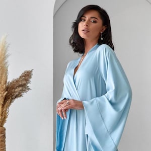 May include: A light blue robe with wide sleeves and a matching belt. The robe is made of a soft, flowing fabric and has a V-neckline. The model is wearing the robe in a studio setting.