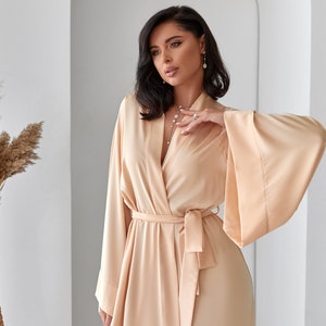 K&ouml;nnte beinhalten: Eine lange, flie&szlig;ende, hellbeige Satinrobe mit V-Ausschnitt und Bindeg&uuml;rtel. Die Robe hat weite &Auml;rmel und eine lockere Passform.