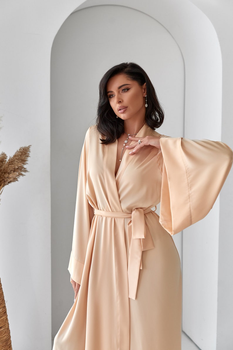 K&ouml;nnte beinhalten: Eine lange, flie&szlig;ende, hellbeige Satinrobe mit V-Ausschnitt und Bindeg&uuml;rtel. Die Robe hat weite &Auml;rmel und eine lockere Passform.
