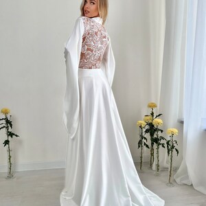 May include: A white satin robe with a floral lace back panel and long, flowing sleeves. The robe has a high collar and a long, elegant skirt. The fabric drapes gracefully, creating a luxurious and romantic look. The robe is suitable for bridal wear or special occasions.