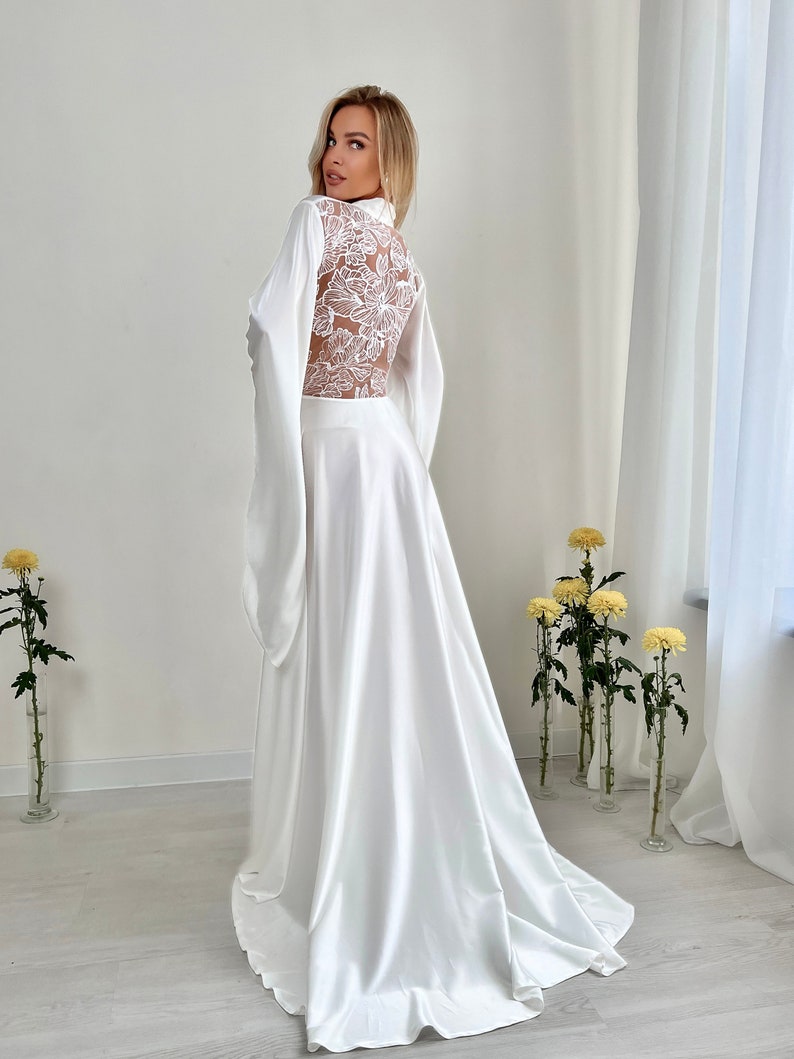 May include: A white satin robe with a floral lace back panel and long, flowing sleeves. The robe has a high collar and a long, elegant skirt. The fabric drapes gracefully, creating a luxurious and romantic look. The robe is suitable for bridal wear or special occasions.