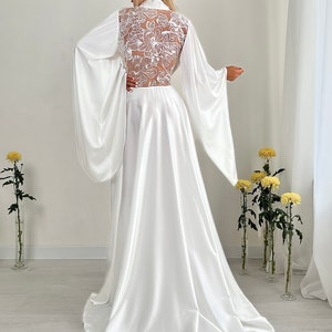 May include: A white satin bridal robe with long, flowing sleeves and a high collar. The back features a sheer panel with intricate floral lace detailing. The robe's skirt flows to the floor, creating an elegant silhouette. The image is taken indoors.