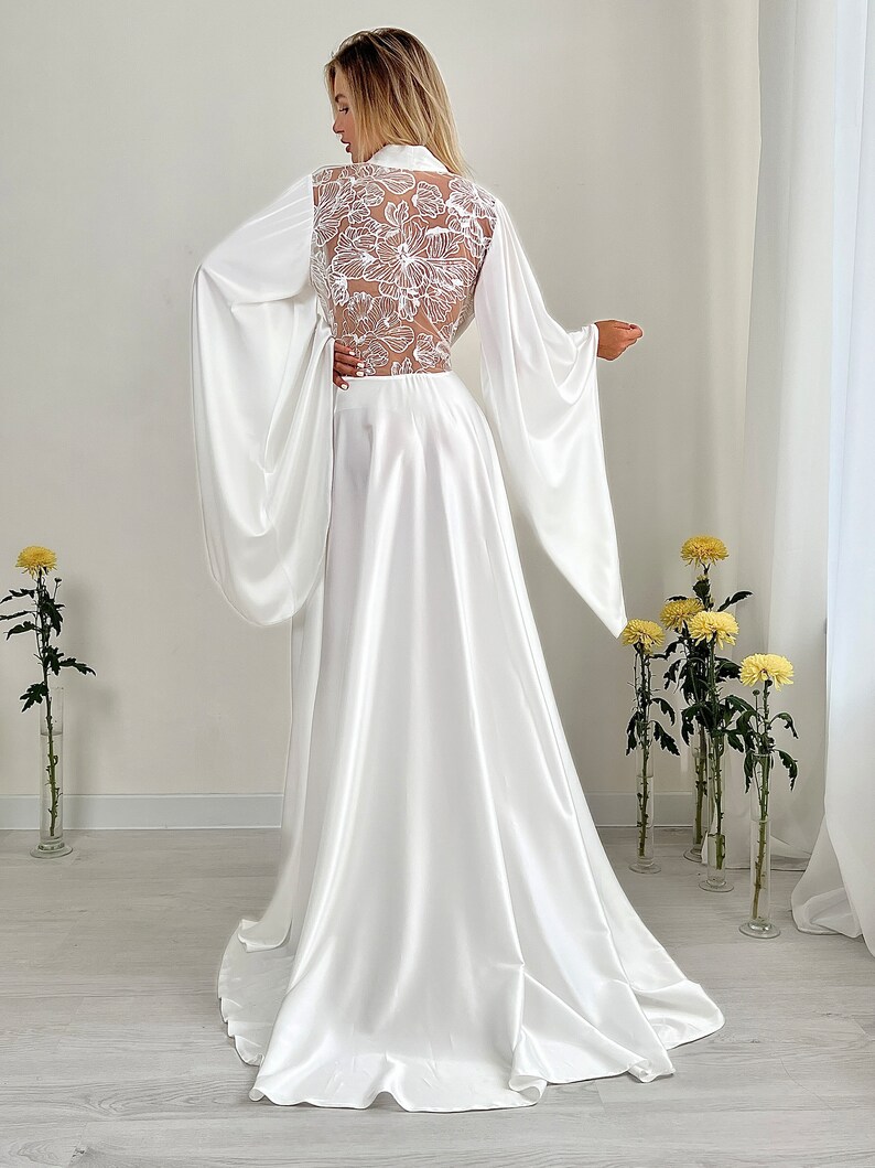 May include: A white satin bridal robe with long, flowing sleeves and a high collar. The back features a sheer panel with intricate floral lace detailing. The robe's skirt flows to the floor, creating an elegant silhouette. The image is taken indoors.