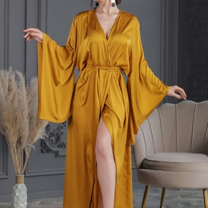 May include: A long, mustard yellow satin robe with a V-neckline, long sleeves, and a tie belt. The robe has a side slit and is worn by a person with bare legs.
