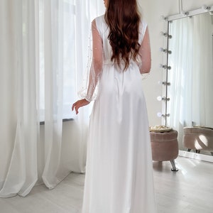 May include: A white satin robe with long sleeves and a pearl-embellished sheer overlay. The robe has a sash tie at the waist and a flowing skirt.