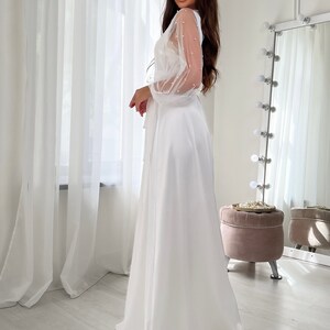 May include: A white satin maxi dress with sheer, pearl-embellished sleeves. The dress has a wrap-style bodice and a flowing skirt.