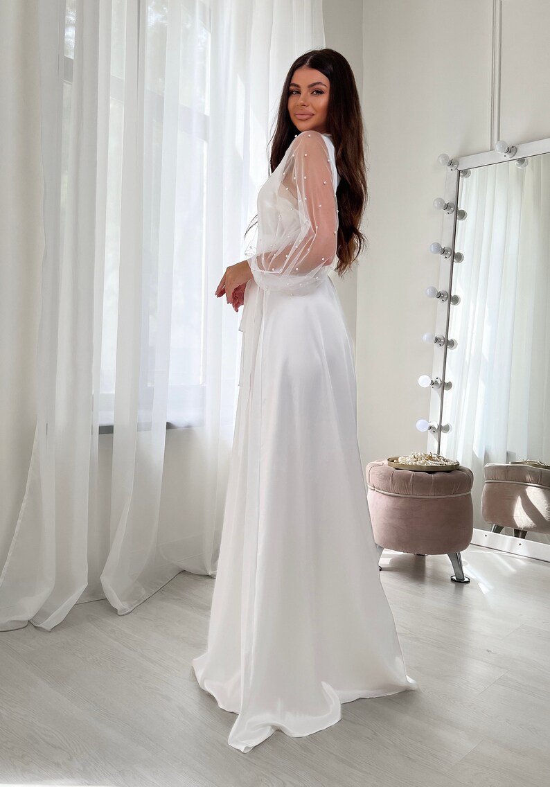 May include: A white satin maxi dress with sheer, pearl-embellished sleeves. The dress has a wrap-style bodice and a flowing skirt.