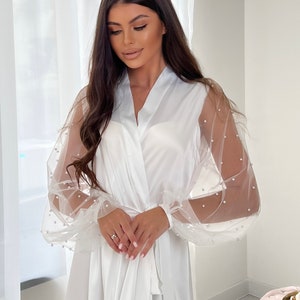 May include: A white satin robe with sheer, pearl-embellished sleeves. The robe has a long, flowing silhouette and a tie belt.