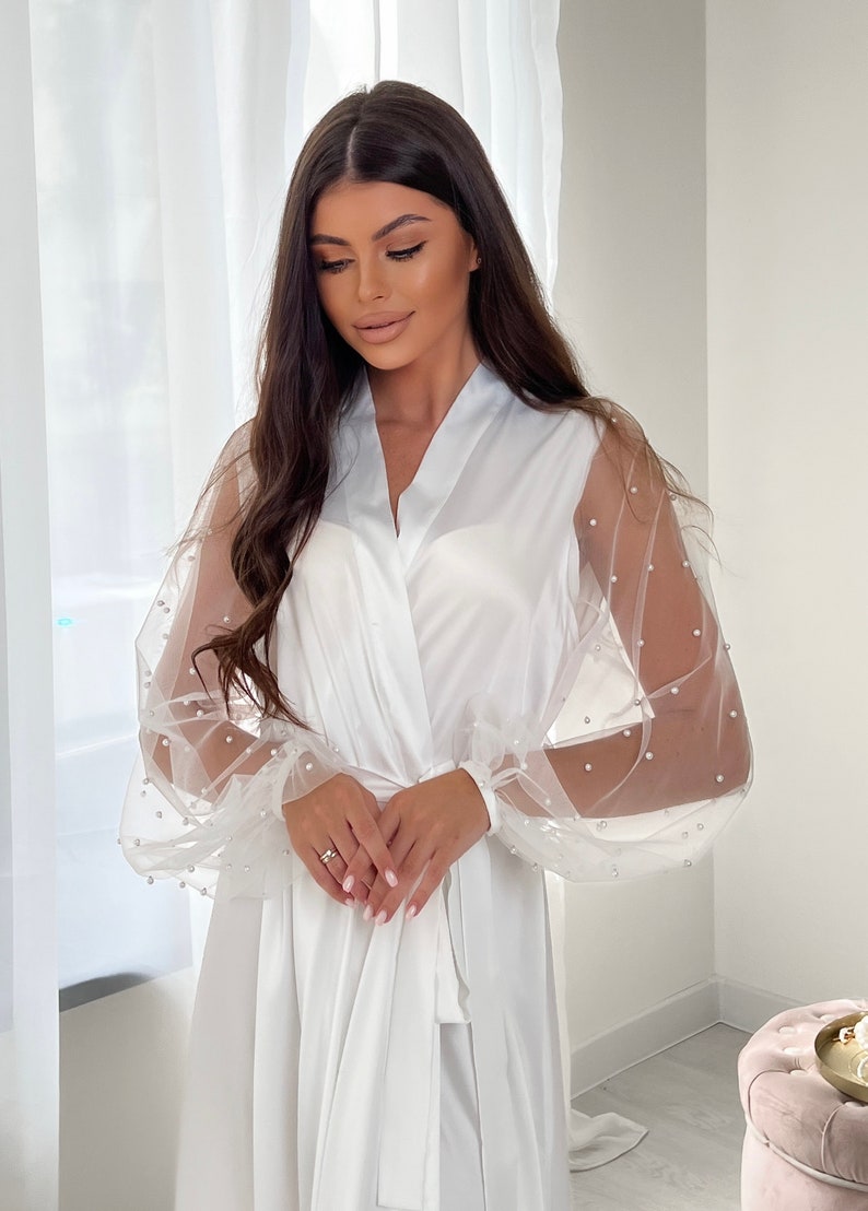 May include: A white satin robe with sheer, pearl-embellished sleeves. The robe has a long, flowing silhouette and a tie belt.
