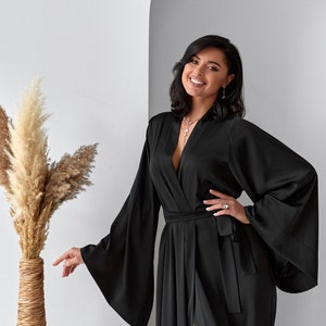 K&ouml;nnte beinhalten: Eine Frau tr&auml;gt eine schwarze Satin-Robe mit langen &Auml;rmeln und einem Bindeg&uuml;rtel. Die Robe hat einen tiefen V-Ausschnitt und einen Schlitz an der Seite. Sie l&auml;chelt und schaut in die Kamera.