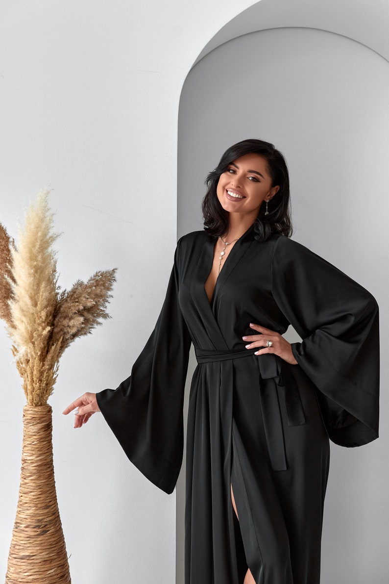 K&ouml;nnte beinhalten: Eine Frau tr&auml;gt eine schwarze Satin-Robe mit langen &Auml;rmeln und einem Bindeg&uuml;rtel. Die Robe hat einen tiefen V-Ausschnitt und einen Schlitz an der Seite. Sie l&auml;chelt und schaut in die Kamera.