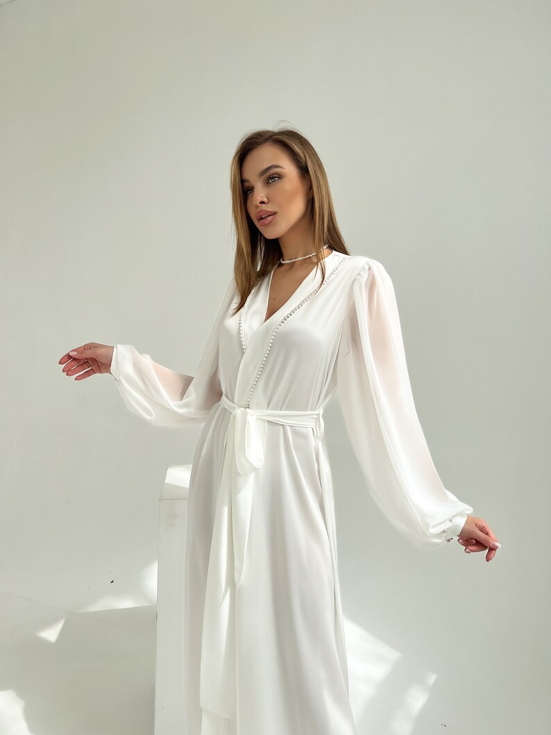 Puede incluir: Una bata de sat&eacute;n blanco con mangas transparentes y cintur&oacute;n a juego. La bata presenta un escote en V con detalles de perlas y pu&ntilde;os abotonados. El dise&ntilde;o largo y fluido es ideal.
