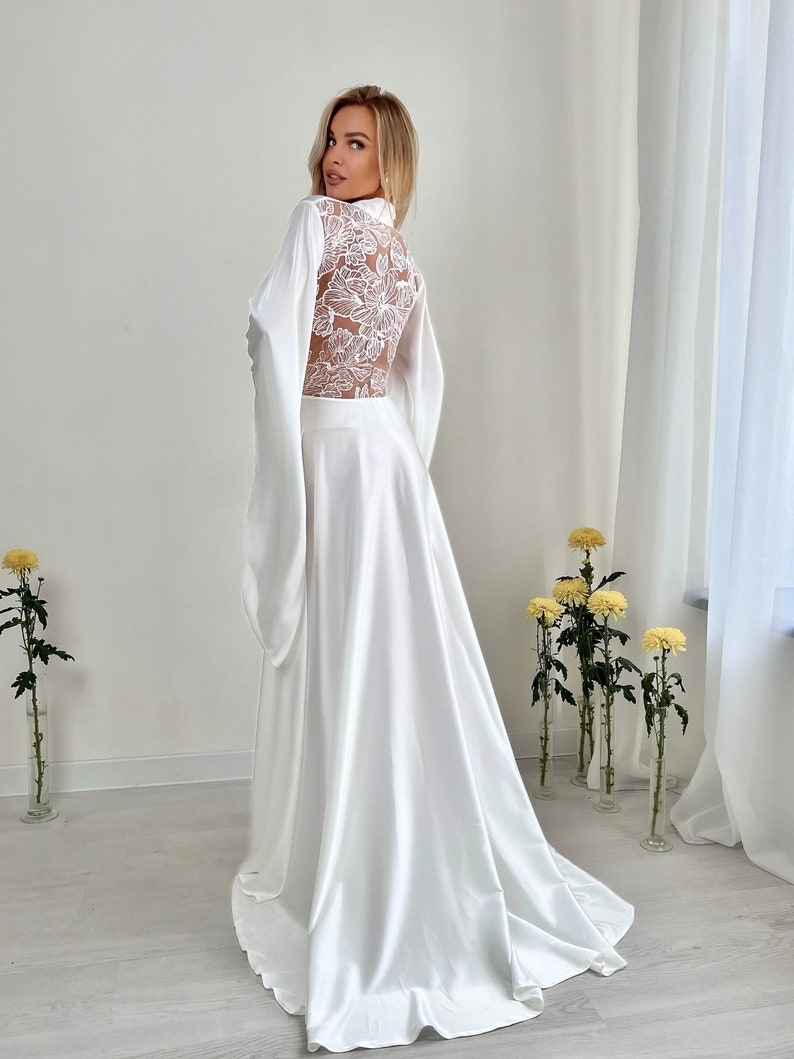 May include: A white satin robe with a floral lace back and long sleeves. The robe has a high collar and a flowing skirt. The model is wearing the robe in a well-lit room with yellow flowers in vases.