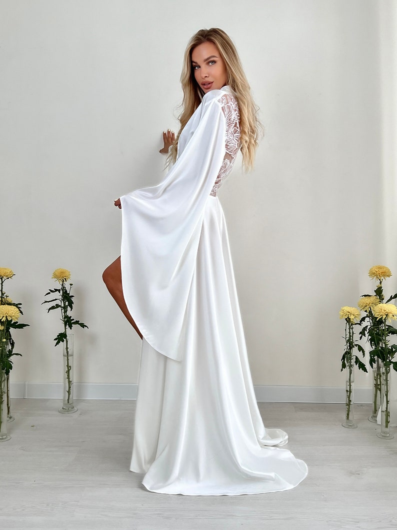 May include: A flowing white satin robe with wide sleeves and a lace back panel. The robe is floor-length and has a high slit, creating an elegant and airy silhouette. The design is perfect for bridal wear or special occasions.