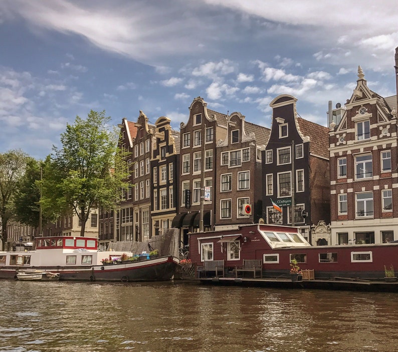 K&ouml;nnte beinhalten: Blick auf einen Kanal in Amsterdam, Niederlande, mit traditionellen Backsteingeb&auml;uden, die das Ufer s&auml;umen. Ein Hausboot liegt im Kanal, mit einer rot-wei&szlig; gestreiften Markise. Die Geb&auml;ude sind eine Mischung aus rotem Backstein und wei&szlig;en Verzierungen, einige mit Giebeld&auml;chern.