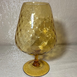 May include: A vintage amber-colored brandy snifter glass. The glass has a wide, rounded bowl with a textured, dimpled pattern and a short stem. The base is a flat, circular foot. The glass is made of a transparent material.