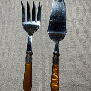 May include: A silver serving fork and cake server set. The utensils have ornate silver detailing where the handles meet the serving ends. The handles are a translucent amber color.