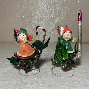 May include: Two vintage Christmas elf figurines. One elf wears an orange coat and green hat, the other wears a green coat and red hat. Both are on holly bases with wire stands. A candy cane and a silver candle complete the festive scene.