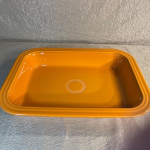 May include: A rectangular orange ceramic baking dish with rounded corners and a wide, flat rim featuring three concentric embossed rings. The interior base of the dish also displays a centered design of three concentric embossed circles.