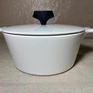 May include: White enamel casserole dish with a black handle on the lid. The dish has a rounded shape with a lid and two side handles. The casserole dish is on a neutral background.