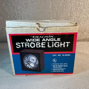 May include: A vintage Realistic Wide Angle Strobe Light in its original box. The box features a black, red, and blue color scheme with the product name prominently displayed. The strobe light itself is visible in a small square window on the box.