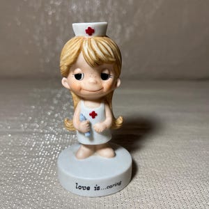 May include: A figurine of a nurse with blonde hair, wearing a white uniform and cap with a red cross. She is holding a blue object and stands on a white base with the text "love is...caring".