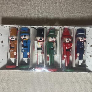 May include: A set of six miniature wooden nutcracker figurines in a clear plastic box. Each nutcracker is painted in a different color, including brown, blue, white, green, red, and navy. The box is decorated with white stars.