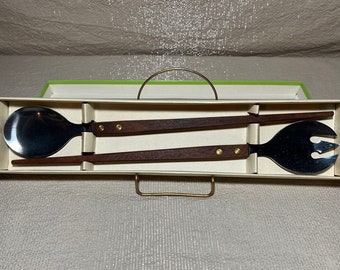 May include: A salad serving set with a spoon and a fork, each with a stainless steel head and a dark wood handle. The handles are attached to the heads with brass rivets. The set is presented in a cream-colored box with a green lid.