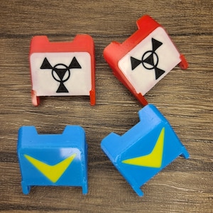 May include: Four plastic game pieces, two red and two blue. The red pieces have a white background with a black radiation symbol. The blue pieces have a yellow chevron shape on a blue background.