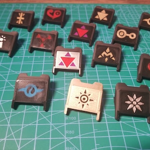 May include: A collection of 13 black plastic keycaps with different colored and white symbols. The symbols include a cross, a heart, a triangle, a circle, a star, a crescent moon, a spiral, and a sun.