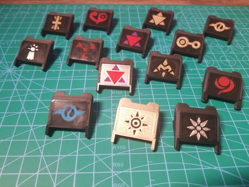 May include: A collection of 13 black plastic keycaps with different colored and white symbols. The symbols include a cross, a heart, a triangle, a circle, a star, a crescent moon, a spiral, and a sun.