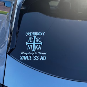 May include: A white decal on a car window displays the word "ORTHODOXY" above a cross symbol and the words "NI KA". Below, it reads "Keeping it Real SINCE 33 AD". The car is a light blue colour.