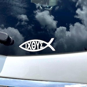 Puede incluir: Una pegatina blanca con el símbolo del pez Ichthys, con la palabra griega "ΙΧΘΥΣ" en su interior, está adherida a la ventana trasera de un vehículo. La pegatina es un símbolo religioso, y el fondo muestra un cielo nublado.