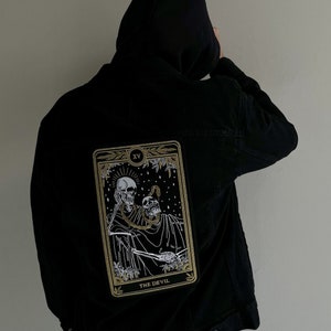 May include: Black denim jacket with a gold and white embroidered tarot card patch on the back. The card is the XV, The Devil.