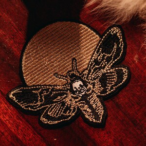 Könnte beinhalten: Gestickter Aufnäher mit einer schwarzen Motte mit Totenkopf-Design, vor einem goldenen Kreis. Der Aufnäher befindet sich auf einer dunklen Holzoberfläche, mit einer fellartigen Textur im Hintergrund. Ein einzigartiges Accessoire.