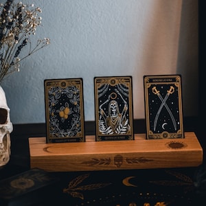 May include: Three tarot cards with gold accents and black backgrounds are displayed on a wooden stand. The cards are titled "Queen of Swords", "Queen of Wands", and "Swords of Oath".