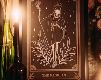 May include: Black framed tarot card print featuring "The Magician" with a skeletal figure holding a star and orb. The card has gold accents and the text "THE MAGICIAN". The image is displayed on a wooden surface.