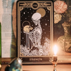 May include: A black tarot card with gold accents depicting the Strength card. The card features a white skeleton holding a skull in its arms. The card is set against a dark background with gold stars and a gold moon. The card is labelled "VIII" and "STRENGTH".