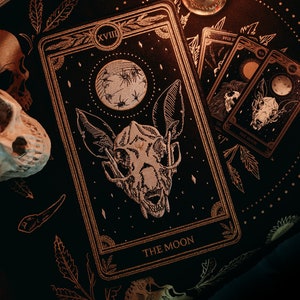 May include: A tarot card reading with a black background and gold accents. The card is the "The Moon" card, which is the 18th card in the Major Arcana. The card features a black and white illustration of a wolf's head with a crescent moon above it. The card is surrounded by other tarot cards, a lit candle, and skulls.