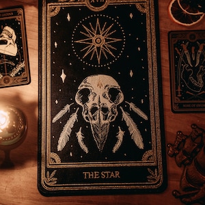 May include: A black tarot card with gold accents depicting a skull with feathers and a star. The card is labelled "The Star" and "XVII".