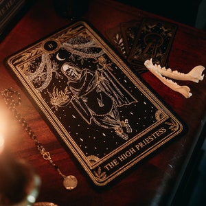 May include: A black tarot card with gold accents depicting a skeletal figure in a hooded robe. The card is labelled "The High Priestess" and features a crescent moon, stars, and a vine-like pattern.