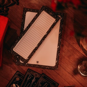 May include: Two black and white patterned notebooks with blank pages. The notebooks are on a wooden surface with a red book, a candle, and tarot cards.