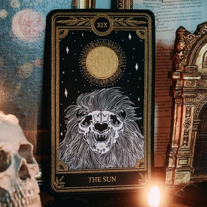 May include: A black tarot card with gold accents depicting the Sun card. The card features a white embroidered image of a lion's head with a sun behind it. The card is labeled "XIX THE SUN".
