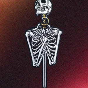 May include: A detailed black and white patch depicting a skeleton with a sword through its rib cage. The skull is at the top, with a gold-coloured neck. The patch is set against a dark red background.